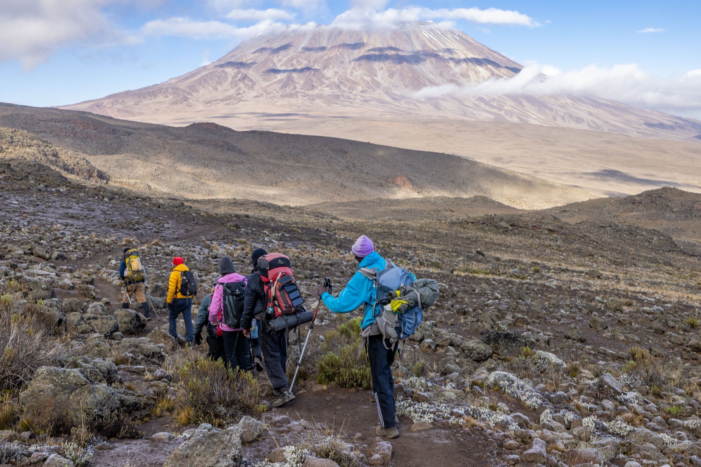 A line of trekkers walking down a rocky trail with Kibo behind them